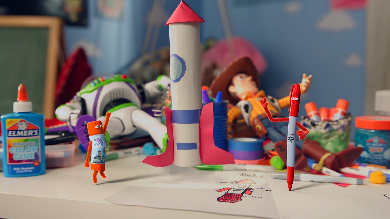 A handmade paper rocket with a pink nose cone standing on a desk, flanked by a bottle of Elmer's Color Glue on the left and a red Sharpie marker with pipe cleaner arms on the right, toy figures behind in soft focus, directed by Rick Wayne